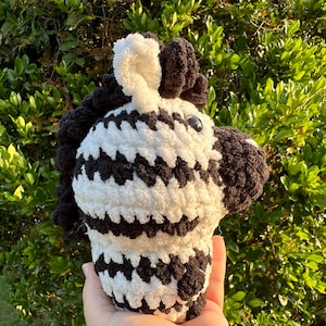 May include: A crocheted zebra plush toy with black and white stripes. The toy has a fluffy texture and is holding a small, black and white striped ball.