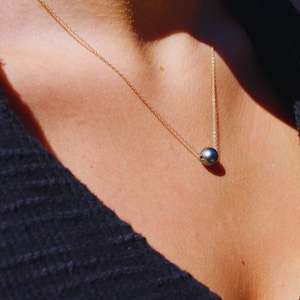 Puede incluir: Un collar de cadena dorada con un único colgante de perla gris oscuro. La perla es redonda y cuelga en el centro del pecho. El collar se lleva sobre una prenda azul oscuro.