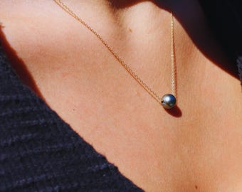 May include: A delicate gold chain necklace featuring a single, smooth, dark grey spherical bead pendant. The necklace rests against the skin, showcasing the contrast between the metallic gold chain and the polished, round, dark grey centerpiece.