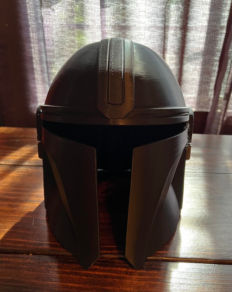 May include: A black Mandalorian helmet, with a rounded top and a central ridge. The helmet has a visor opening and angular cheek plates. The matte-finish helmet is displayed on a wooden surface.
