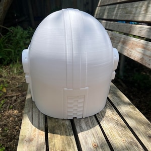 May include: A white 3D-printed helmet with a rounded top and rectangular features. The helmet has two side protrusions and a textured design on the back. It is sitting on a wooden bench.