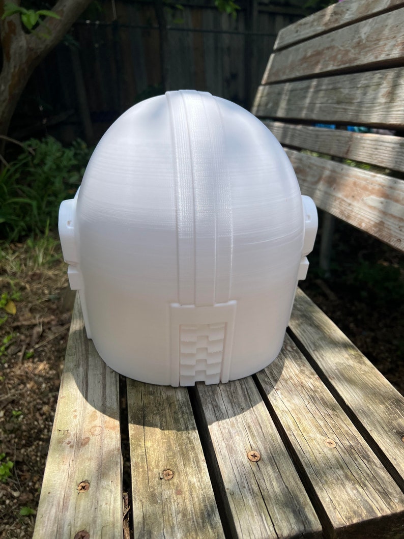 May include: A white 3D-printed helmet with a rounded top and rectangular features. The helmet has two side protrusions and a textured design on the back. It is sitting on a wooden bench.