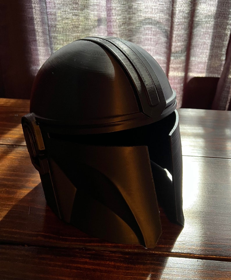 May include: A black Mandalorian helmet, with a rounded crown and a visor. The helmet has a raised central ridge and is made of a matte material. It is displayed on a wooden surface. The design is inspired by the Star Wars series.