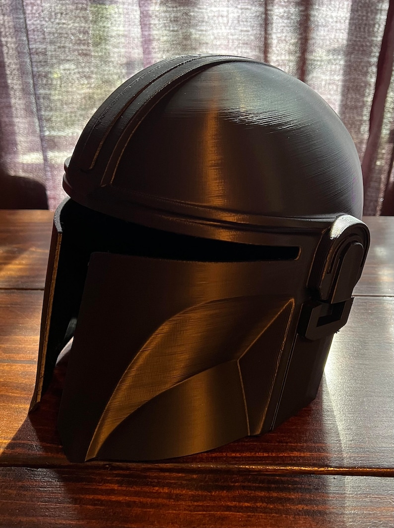 May include: A black Mandalorian helmet, featuring a rounded crown and angular details. The helmet includes a visor and intricate design, possibly 3D-printed. The dark hue and textured finish give it a robust look, ideal for cosplay or display.