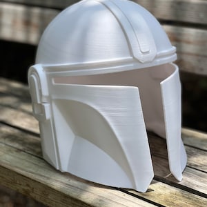 May include: A white Mandalorian helmet with a T-shaped visor and a distinctive angular design. The helmet is made of a solid, opaque material and has a smooth, matte finish. The helmet is sitting on a wooden surface.
