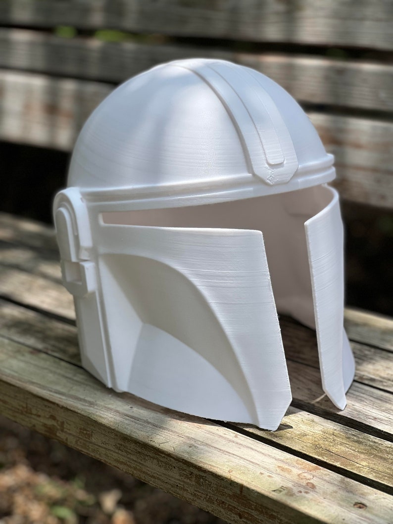 May include: A white Mandalorian helmet with a T-shaped visor and a distinctive angular design. The helmet is made of a solid, opaque material and has a smooth, matte finish. The helmet is sitting on a wooden surface.