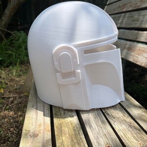 May include: A white Mandalorian helmet, placed on a wooden bench. The helmet features a rounded crown, a visor, and unique side elements. The surface shows layer lines from 3D printing. This is a prop or costume piece.