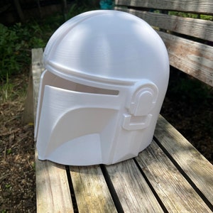 May include: A white Mandalorian helmet with a rounded crown and a distinctive visor. The helmet has detailed side accents and a smooth finish. It is placed on a weathered wooden surface, likely a prop or collectible item.
