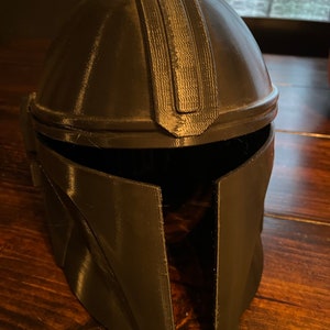 May include: A dark grey Mandalorian helmet, possibly 3D-printed, with a rounded crown and angular cheek plates. The helmet has a textured surface and a visor opening. The design is inspired by a popular science fiction character.