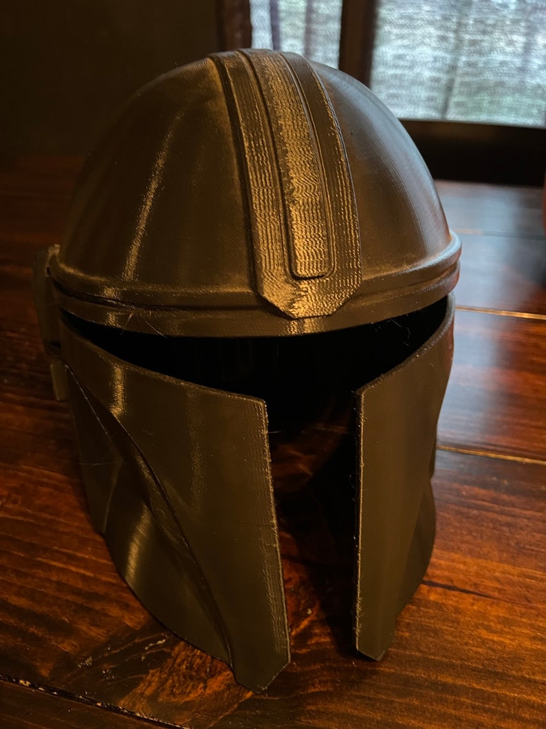 May include: A dark grey Mandalorian helmet, possibly 3D-printed, with a rounded crown and angular cheek plates. The helmet has a textured surface and a visor opening. The design is inspired by a popular science fiction character.