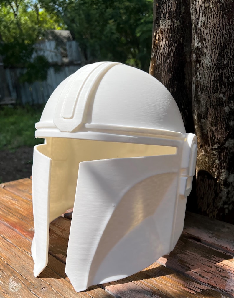 May include: A white Mandalorian helmet, with a rounded crown and angular cheek plates. The helmet has a raised central ridge and is made of a solid, opaque material. It is displayed on a wooden surface, with a natural background.
