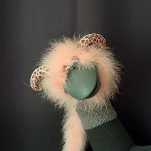 May include: A hand puppet featuring a fluffy light pink body with long synthetic fur, two rounded ears with a brown and black leopard print pattern, two small plastic eyes, and a solid dark gray beak-like mouth structure extending forward.