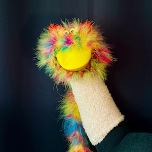 May include: A hand puppet featuring a creature with long, multi-colored faux fur in shades of pink, yellow, blue, and green. It has two googly eyes, red ears, and a large, smooth, solid yellow circular mouth. The lower section is a white textured fabric.