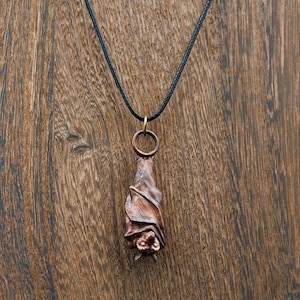 May include: A copper-coloured bat pendant hangs from a black cord necklace. The bat is sculpted in a realistic style with wings folded and a detailed face.