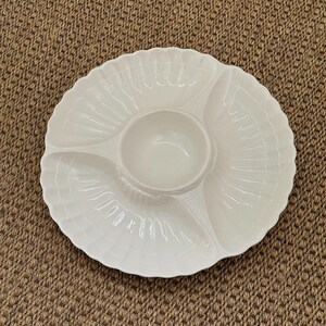 May include: A white serving platter with a central dip bowl and three surrounding sections. The platter has a scalloped edge and a shell-like pattern. Suitable for serving snacks or appetizers at a gathering.