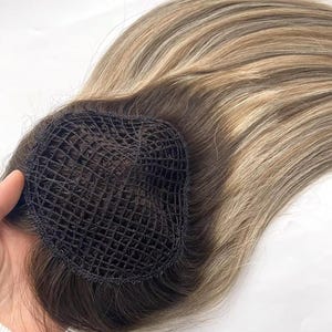 May include: A hairpiece featuring a dark brown mesh base and light brown and blonde highlights. The hairpiece is designed to add volume and length to natural hair. The hair is styled in loose waves.