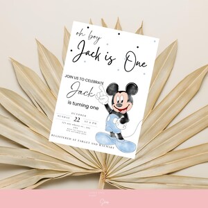 以下が含まれることがあります： 「oh boy Jack is One」のテキストとミッキーマウスの漫画の画像が描かれた白い招待状。 招待状には、日付と時間を含む1歳の誕生日のお祝いの詳細が含まれています。 カードは乾燥したヤシの葉を背景に設定されています。