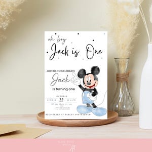 以下が含まれることがあります： ミッキーマウスの誕生日招待状。「oh boy, Jack is One」と書かれ、お祝いの詳細が記載されています。デザインには、青、黒、白の漫画ミッキーマウスのイラストが白い背景に描かれています。