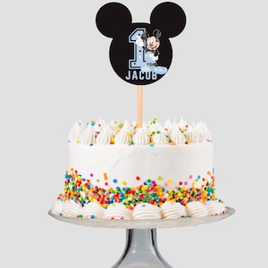 May include: A white frosted cake with colourful sprinkles and white frosting swirls. A Mickey Mouse-themed cake topper with the number 1 and the name "Jacob" is placed on top. The cake is on a clear glass cake stand.