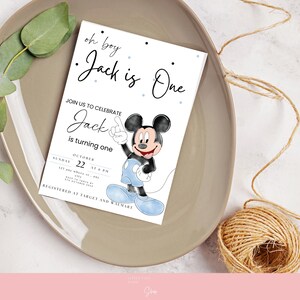 以下が含まれることがあります： 「oh boy Jack is One」の文字とミッキーマウスのイラストが描かれた白い招待状。招待状は、緑と紐の玉が添えられたベージュの皿の上にあります。「Jack is turning one」の文字も表示されています。