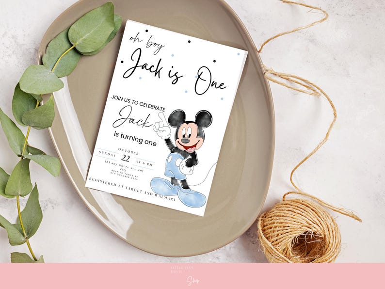 以下が含まれることがあります： 「oh boy Jack is One」の文字とミッキーマウスのイラストが描かれた白い招待状。招待状は、緑と紐の玉が添えられたベージュの皿の上にあります。「Jack is turning one」の文字も表示されています。
