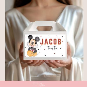 May include: White gable box with a handle, featuring a Mickey Mouse illustration and the name "JACOB" in red letters. The text "Turns Two" is also included. Black and red polka dots decorate the box.