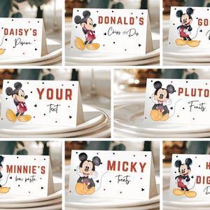 May include: Set of Mickey Mouse-themed table cards with various food and drink labels. Each card features a Mickey Mouse illustration and text like "Daisy's Popcorn" or "Goofy's Drinks" in red and black. The cards are on white plates.