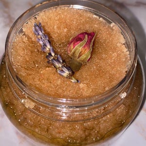 Op de afbeelding: Een heldere glazen pot gevuld met een bruine, korrelige scrub. Een takje lavendel en een kleine, roze rozenknop liggen bovenop de scrub. De pot staat op een wit oppervlak.