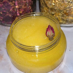 Op de afbeelding: Een heldere pot gevuld met een heldergele, getextureerde scrub. Een kleine, gedroogde roze rozenknop rust in het midden. Potten met gedroogde kruiden en bloemen zijn zichtbaar op de achtergrond, wat wijst op een natuurlijk huidverzorgingsproduct.