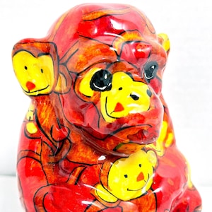 May include: A red ceramic monkey figurine with yellow and black details. The monkey is holding a smaller monkey figurine in its arms.