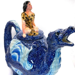 Op de afbeelding: Een blauwe en witte keramische theepot in de vorm van een draak met een vrouw in een luipaardprintjurk die op zijn rug rijdt.