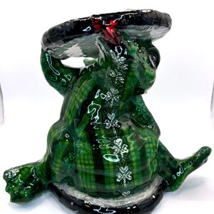May include: A green ceramic frog figurine with a plaid pattern and silver glitter accents. The frog is holding a black and red plate over its head.