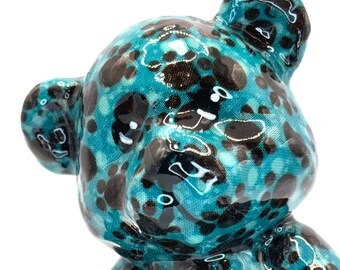 May include: A teal blue ceramic teddy bear with black paw print patterns. The bear has a glossy finish and is sitting with its head tilted.