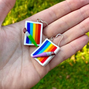 May include: Handmade earrings with a rainbow design and a miniature paintbrush. The earrings are rectangular with a white border and a colourful rainbow pattern. The paintbrushes have green and purple handles.