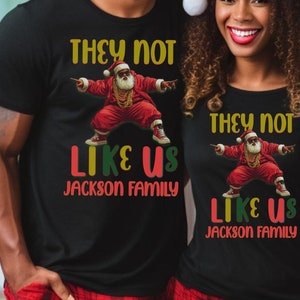 Puede incluir: Una pareja lleva camisetas negras a juego con un gr&aacute;fico de un Pap&aacute; Noel bailando y el texto "They not like us Jackson Family". Llevan pantalones de pijama de cuadros rojos y negros y gorros de Pap&aacute; Noel.