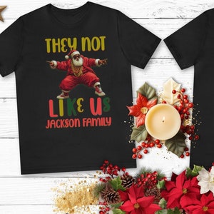 Puede incluir: Un conjunto de cuatro camisetas negras con un gr&aacute;fico de Pap&aacute; Noel rapeando. Las camisetas tienen el texto "They Not Like Us Jackson Family" impreso en ellas.