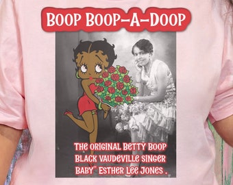 Puede incluir: Una camiseta rosa claro con un gráfico de Betty Boop sosteniendo rosas, con el texto "BOOP BOOP-A-DOOP" arriba y "THE ORIGINAL BETTY BOOP BLACK VAUDEVILLE SINGER BABY" ESTHER LEE JONES" abajo. La camiseta es usada por una persona.