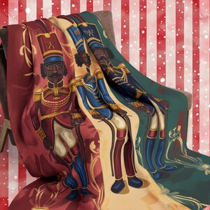 May include: A red, green, and gold throw blanket with a whimsical design featuring three toy soldiers. The soldiers are dressed in red, blue, and green uniforms and have black faces. The blanket has a striped background with white snowflakes.