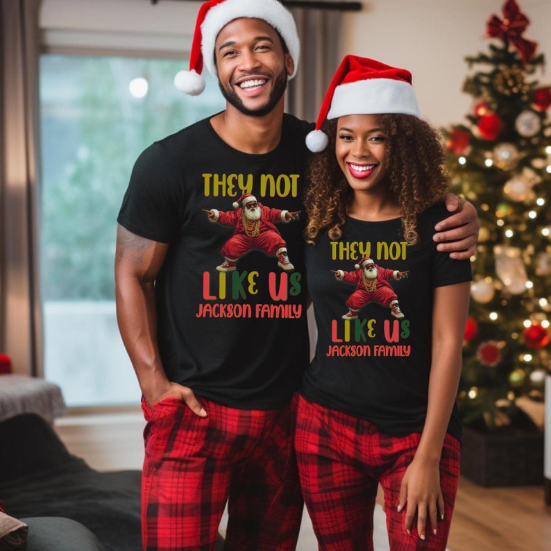 Puede incluir: Una pareja lleva camisetas negras a juego con un gr&aacute;fico de un Pap&aacute; Noel bailando y el texto "They not like us Jackson Family". Llevan pantalones de pijama de cuadros rojos y negros y gorros de Pap&aacute; Noel.
