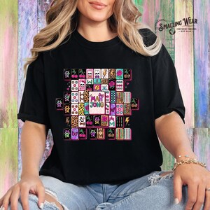 May include: A black t-shirt featuring a central graphic of various mahjong tiles arranged in a cluster. The tiles display diverse patterns including cherries, skulls, leopard print, roses, lightning bolts, and the text "MAH JONG" in the center.