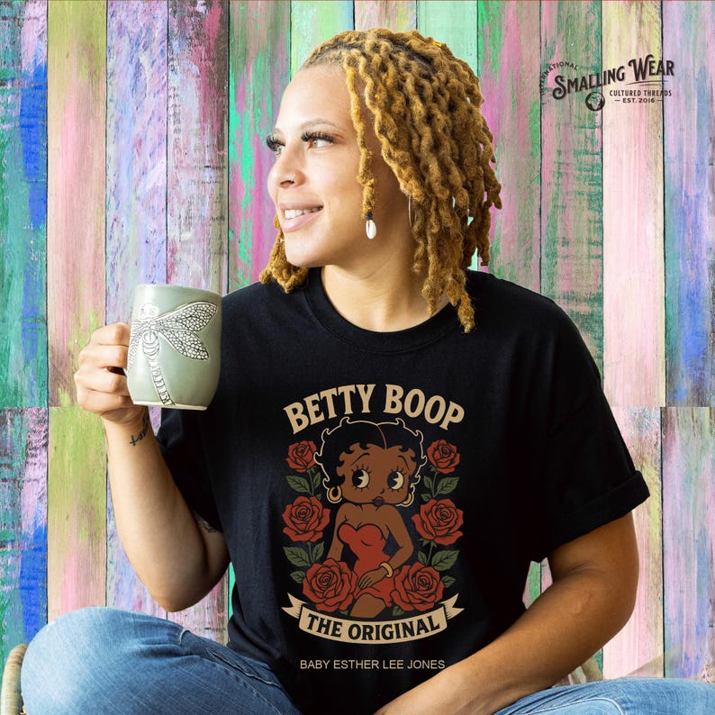 Puede incluir: Camiseta negra con un gr&aacute;fico de Betty Boop, rosas rojas y el texto "BETTY BOOP THE ORIGINAL". La camiseta se combina con unos vaqueros azules y una taza verde con un dise&ntilde;o de lib&eacute;lula.