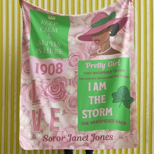 Op de afbeelding: Roze en groene deken met een bloemenpatroon en de tekst "Keep Calm A Alpha Is Here 1908" en "Pretty Girl They Whispered To Her You Can Not Withstand The Storm I Am The Storm She Whispered Back Soror Janet Jones".