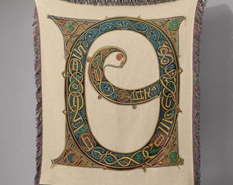 May include: A beige throw blanket with a large, ornate letter "G" in the center. The letter is detailed with blue, green, and gold Celtic knotwork. The blanket has a fringed edge.