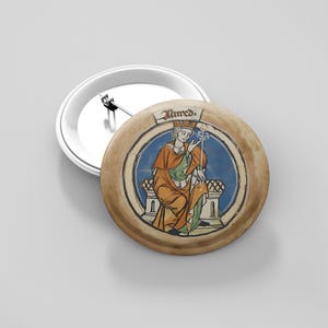 May include: A round button pin featuring a medieval illustration of a figure in orange and green robes, seated on a throne. The figure holds a staff, and the word "Alfred" is written above. The pin has a white metal backing.
