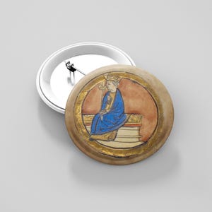 May include: A circular button badge with a medieval illustration of a seated figure in a blue robe and gold crown. The illustration is set within a gold border against a reddish-brown background. The badge has a silver backing.