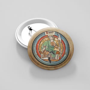 May include: A round button pin featuring a medieval illustration of a seated figure in green robes, holding a staff. The illustration is framed by a blue and red border, with the name "Knur" at the top. The pin has a silver backing.