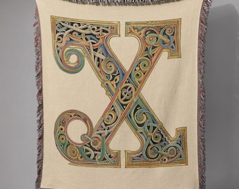 May include: A beige throw blanket featuring a large, ornate letter "X" in a colorful, Celtic-inspired design. The letter is filled with intricate patterns in shades of blue, green, yellow, and red, outlined in gold. The blanket has a fringed edge.