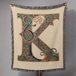 May include: A beige throw blanket featuring a large, ornate letter "K" in a colorful, Celtic-inspired design. The letter is filled with intricate patterns in shades of blue, green, red, and gold. The blanket has a fringed edge.