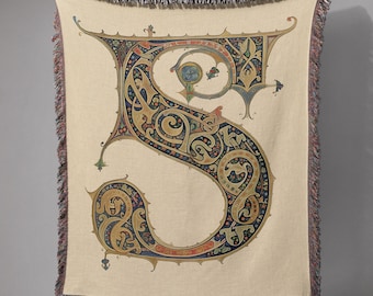 May include: A beige throw blanket featuring a large, ornate letter "S" in a medieval illuminated manuscript style. The letter is filled with intricate patterns in shades of gold, blue, red, and green. The blanket has a fringed edge.