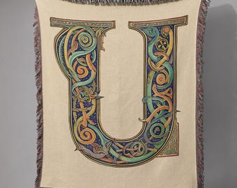 May include: A beige throw blanket with a large, ornate letter "U" in the center. The letter is filled with intricate Celtic knotwork in shades of green, blue, orange, and yellow. The blanket has a fringed edge.